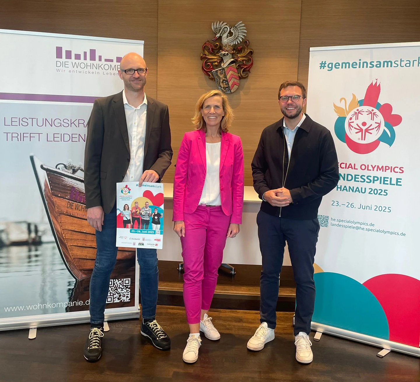 3 Personen stehen lächelnd vor 2 Bannern mit DIE WOHNKOMPANIE Motiv und Special Olympics Motiv,